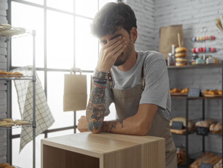 Young hispanic man with beard and tattoos looking distressed in an indoor bakery filled with bread and pastries.